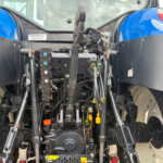 Rear tractor hitch area with hydraulic valves, levers, and hoses; blue fenders on both sides and warning labels nearby