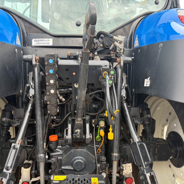 Rear tractor hitch area with hydraulic valves, levers, and hoses; blue fenders on both sides and warning labels nearby