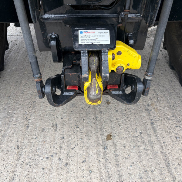 Close-up of a yellow tractor hitch and metal linkage under the rear frame of a dark tractor chassis.