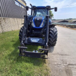 Blue agricultural tractor with front loader facing the camera, parked beside a stone wall and industrial buildings under a blue sky.