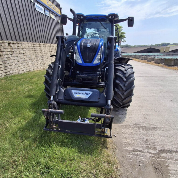 Blue agricultural tractor with front loader facing the camera, parked beside a stone wall and industrial buildings under a blue sky.