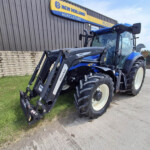 Blue New Holland tractor with front loader parked beside a brick wall and a New Holland Agriculture sign in the background