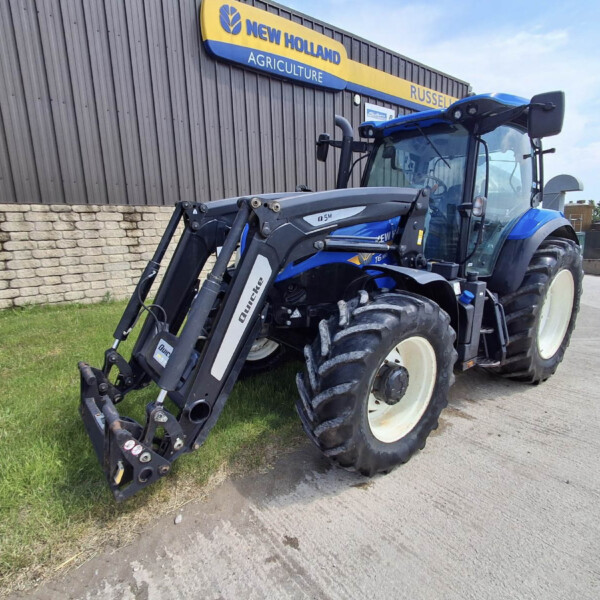 Blue New Holland tractor with front loader parked beside a brick wall and a New Holland Agriculture sign in the background