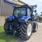 Blue tractor in a parking lot beside a brick wall and agricultural building signage reading Holland Agriculture, large front tires visible