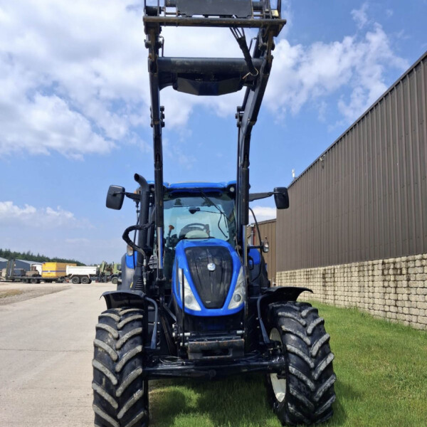 Front view of a blue tractor with its hydraulic loader arms raised, parked beside a metal building on a sunny day.