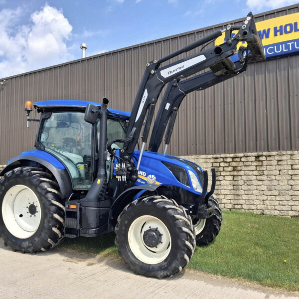 Blue New Holland tractor with a front-end loader raised, parked along a gray metal wall with a New Holland Agriculture sign.