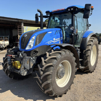 Blue New Holland tractor parked on a dirt lot beside a wooden shed with stacked firewood in the background.
