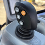 Close-up of a black joystick with circular gray buttons and an orange button featuring a tractor icon on a cabin console.