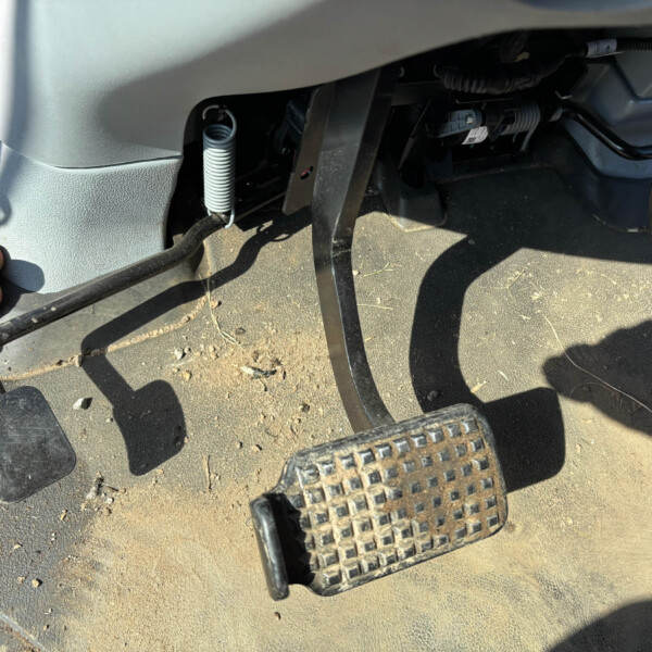 Close-up of a vehicle’s driver footwell showing the accelerator and brake pedals with a dusty floor and a coiled spring nearby.