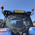 Rear of a blue construction vehicle with two orange beacon lights and license plate FN75 VJJ against a clear blue sky and brick buildings in the background