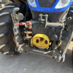 Front view of a blue tractor showing large tread tires, yellow wheel hub, and visible hydraulic hoses and hitch components.