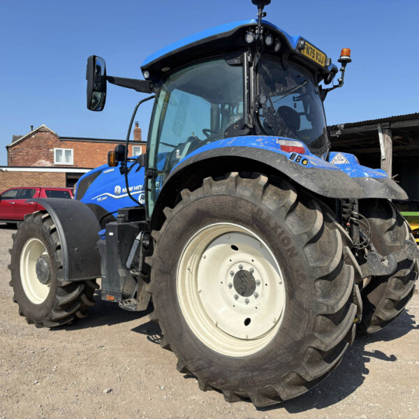 Blue tractor with oversized front tire in a gravel lot, farm buildings visible in the background.