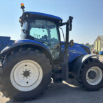 Blue New Holland tractor seen from the front-left with a large front tire in the foreground and an orange beacon on top, in a farmyard.