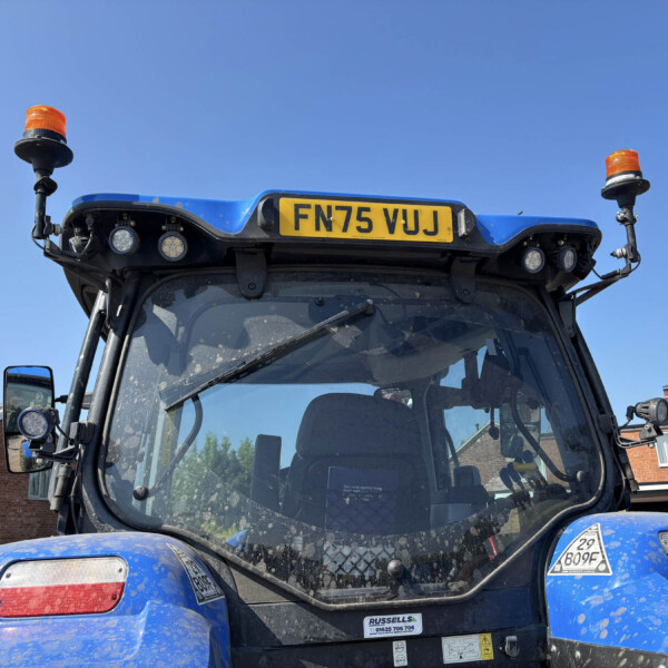 Rear of a blue construction vehicle with two orange beacon lights and license plate FN75 VJJ against a clear blue sky and brick buildings in the background