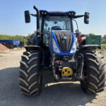Front view of a blue tractor with large chunky tires in a rural equipment yard under a clear sky, with farming gear in the background