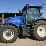 Blue New Holland tractor viewed from the side, showing large front and rear tires and the cab under a clear blue sky.