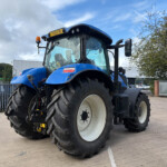 Blue agricultural tractor with oversized rear tires parked in a paved lot, fence and buildings in the background under a partly cloudy sky.
