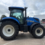 Blue New Holland tractor seen from the side in a fenced industrial lot with large white wheels and big tires.