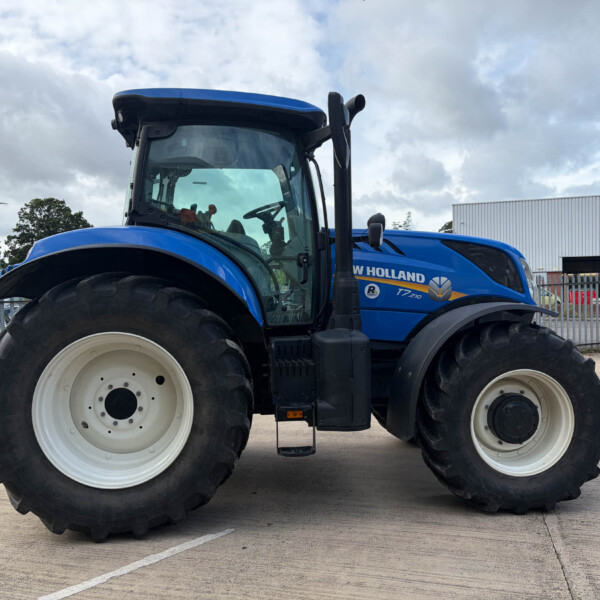 Blue New Holland tractor seen from the side in a fenced industrial lot with large white wheels and big tires.