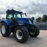 Blue New Holland tractor parked in a lot with a metal fence and cloudy sky behind it.