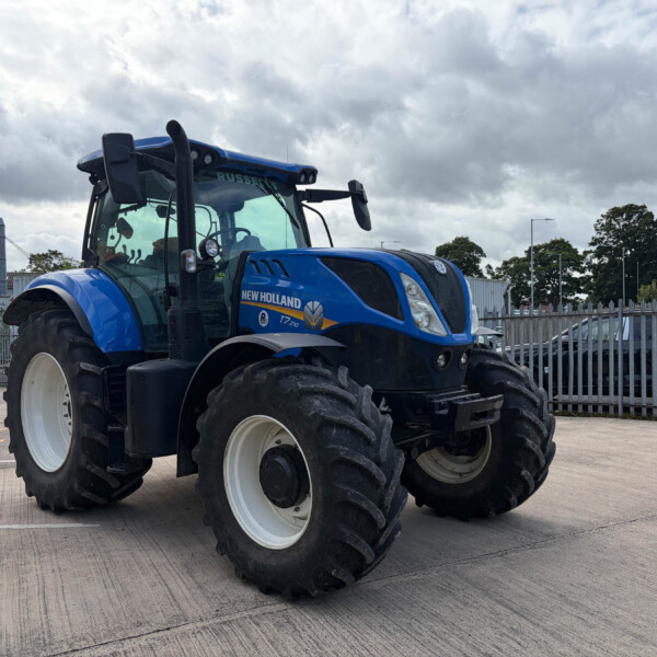 Blue New Holland tractor parked in a lot with a metal fence and cloudy sky behind it.