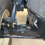 Close-up of a trailer hitch area between a wheel and chassis, showing bolts, cables, and metal brackets.