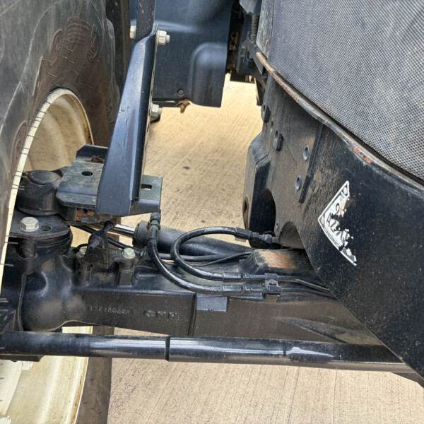 Close-up of a trailer hitch area between a wheel and chassis, showing bolts, cables, and metal brackets.