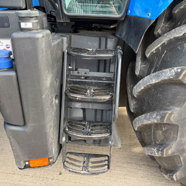 Close-up of a farm tractor step ladder between the large rear tire and a black maintenance panel on a blue tractor surface.
