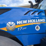 Close-up of a blue New Holland tractor hood with three black vents and part of the 'NEW HOLLAND' logo visible, under a clear sky.