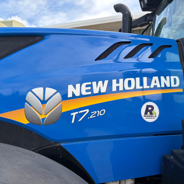 Close-up of a blue New Holland tractor hood with three black vents and part of the 'NEW HOLLAND' logo visible, under a clear sky.