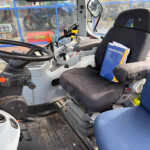 Interior of a tractor cab with steering wheel, controls, and a blue operator's manual on the driver's seat near a blue cloth seat.