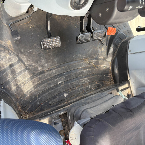 Inside a vehicle cabin showing three pedals (clutch, brake, accelerator) on a dusty floor, with a nearby gear lever and driver-side footwell visible.