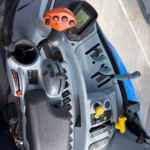 Cockpit of a tractor with an orange multi-button joystick, digital display, steering wheel, and various control levers nearby.