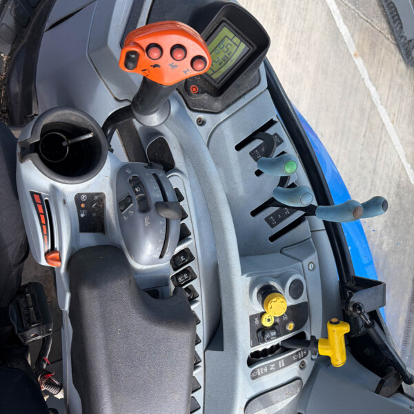 Cockpit of a tractor with an orange multi-button joystick, digital display, steering wheel, and various control levers nearby.