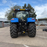 Rear view of a blue tractor with oversized tires in an industrial lot, fence and buildings in the background.