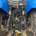 Close-up of a tractor's rear three-point hitch area with hydraulic levers, linkages, and hoses between large dusty tires