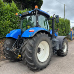 Blue agricultural tractor parked alongside a hedge, with oversized rear tires and orange warning lights on top; a brick building and street in the background.