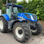 Blue New Holland tractor with oversized rear tires parked beside a green hedge in a yard.