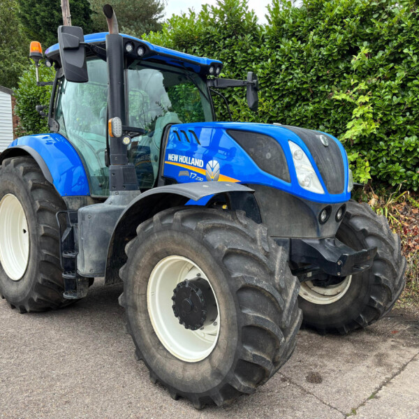 Blue New Holland tractor with oversized rear tires parked beside a green hedge in a yard.