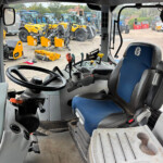 Interior view of a tractor cab with steering wheel, pedals, and blue seat, looking out at a yard full of yellow and blue tractors outside.