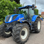 Blue New Holland T7 tractor parked beside a tall green hedge on a paved street, with a red brick building in the background under a cloudy sky.
