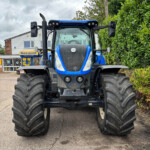 Front view of a blue New Holland agricultural tractor with large tires in a parking lot near a hedge and a building.