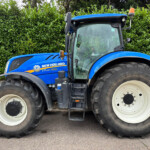 Blue New Holland T7.230 tractor parked beside a dense green hedge, showing oversized wheels at the front and back.