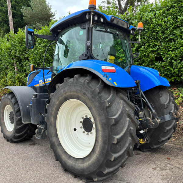 Blue agricultural tractor with large white wheels, cab windows, and orange beacon lights parked beside a green hedge in a driveway edge.
