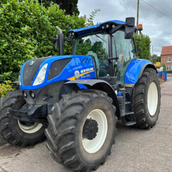 Blue New Holland T7 tractor parked beside a tall green hedge on a paved street, with a red brick building in the background under a cloudy sky.