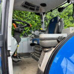Interior view of a New Holland tractor cab with steering wheel, gray control console, and blue/gray seat; door open and green hedge visible outside, floor mat reads 'New Holland'.