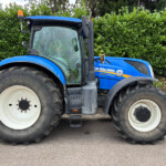 Blue New Holland T7.230 tractor viewed from the side, with large tires and orange beacon, against a hedge background.