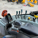 Inside the operator cab of a heavy construction machine, with multiple control levers and a cup holder, yellow equipment visible outside.