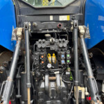 Close-up of a blue tractor's front machinery with hydraulic valves, hoses, and control levers visible at the center.