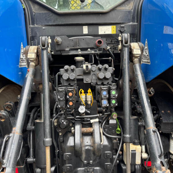 Close-up of a blue tractor's front machinery with hydraulic valves, hoses, and control levers visible at the center.